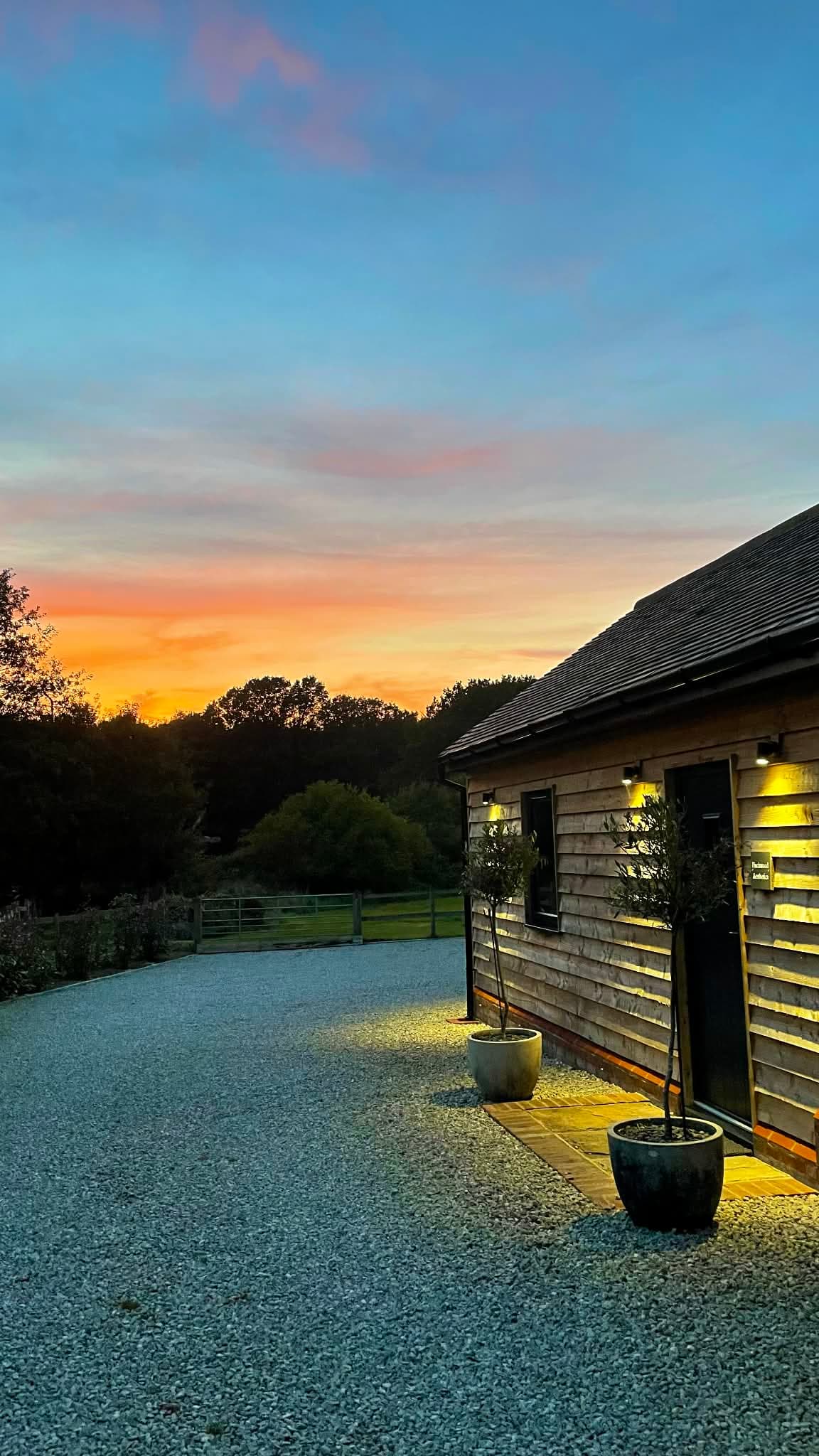 Sunset over the Nurture Body Therapy treatment room in Ashford, Kent — timber-clad building lit by olive-tree lanterns against a vivid Kent sky.